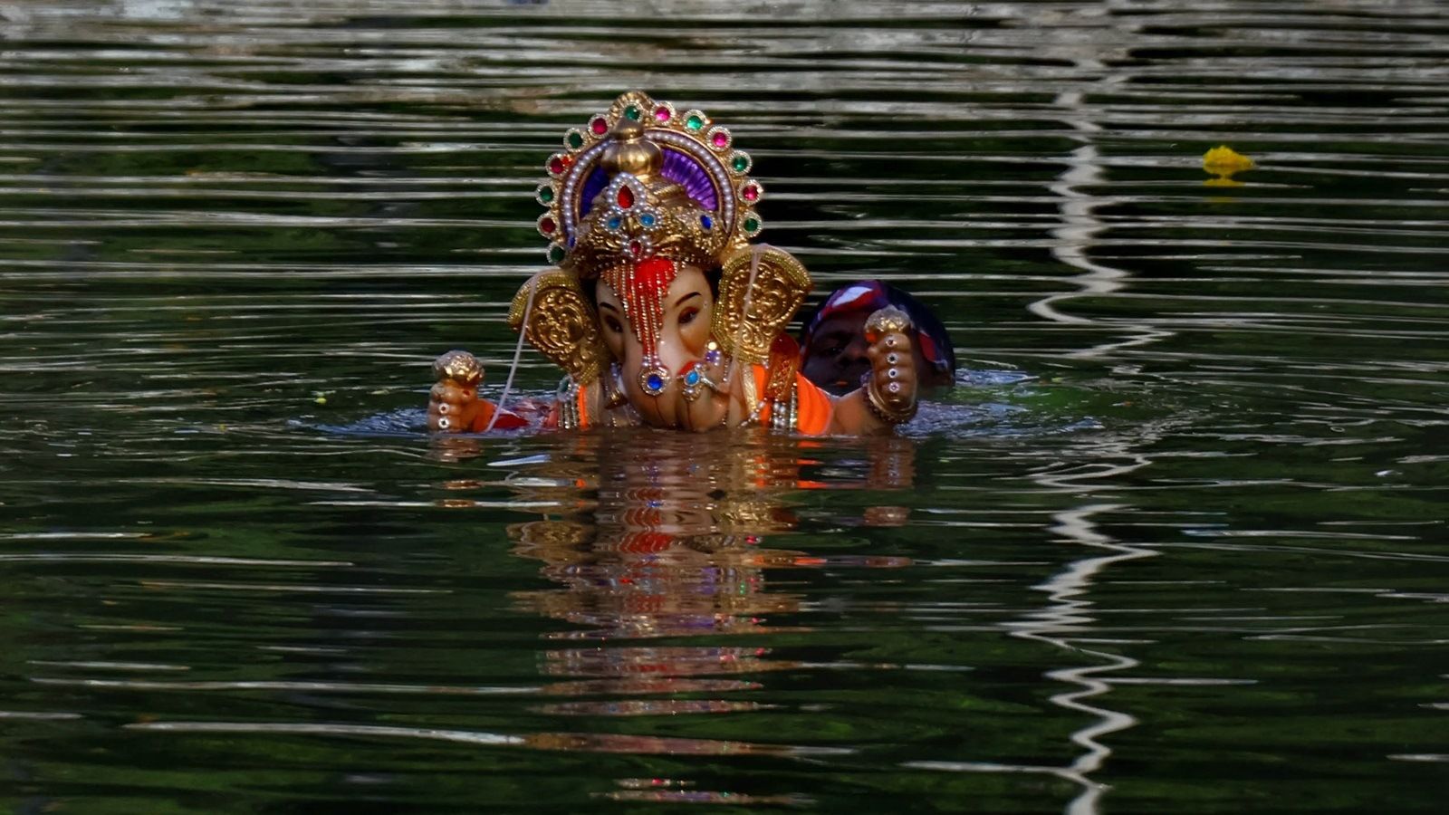 "A beautifully decorated idol of Lord Ganesha being immersed in a body of water during the Ganesh Visarjan ritual. The idol is adorned with colorful jewels and a golden crown, while the water reflects its intricate details. A devotee, partially submerged, is seen assisting in the immersion process."