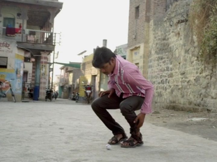  A young boy wearing a pink shirt and black pants bends down to pick up something from the ground in a narrow street of a rural or semi-urban area, surrounded by old houses and parked motorcycles.
