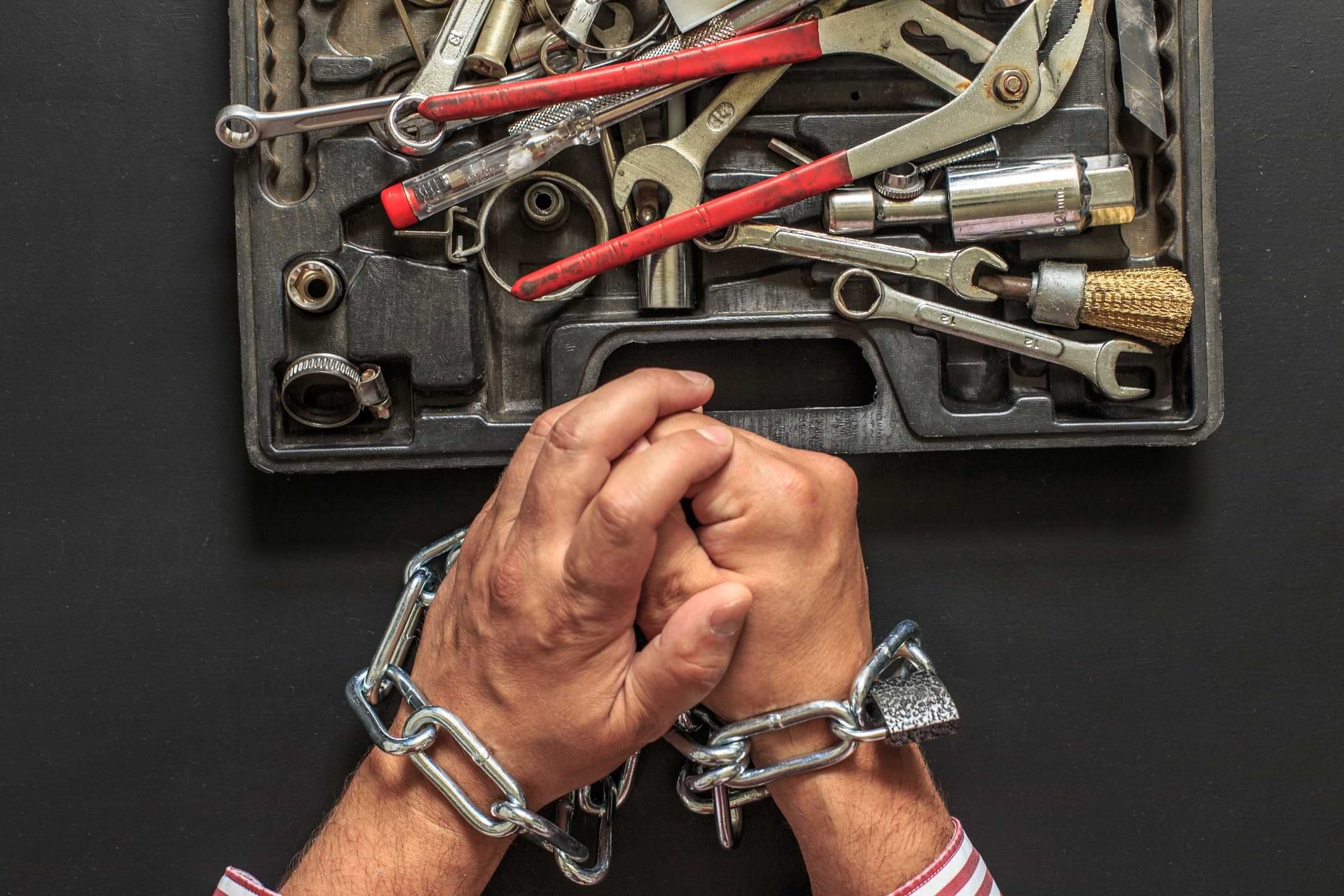 "A pair of human hands tightly bound with metal chains and a padlock, resting on a dark surface. Above the hands is a toolbox filled with various hand tools, including wrenches, pliers, screwdrivers, and sockets. The image conveys a strong sense of restriction, oppression, or forced labor."