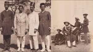 The image appears to be a historical photograph showing a group of men in traditional and colonial-era attire. The left side of the image features men dressed in formal coats, white pants, and traditional Indian headgear, while the right side shows men in turbans, possibly armed, sitting or standing in a posed setting. The photograph seems to depict a moment from colonial-era India, possibly representing different social or professional groups from that time.