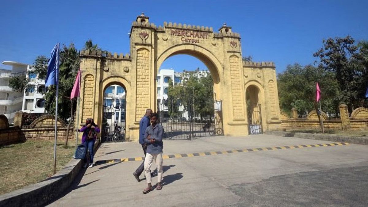A historic yellow stone archway entrance labeled "MERCHANT Campus" stands prominently under a clear blue sky. The gate features decorative battlements and ornamental details. In the foreground, two men walk past the entrance, one using a mobile phone. Another person, carrying a bag, stands near the gate. Behind the archway, modern white buildings and greenery are visible, creating a contrast between old and new architecture. Colorful flags are placed on poles near the entrance.