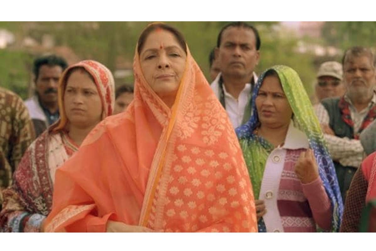 This image appears to show a group of Indian rural women, with a central woman wearing an orange saree and a confident expression. The background consists of other villagers, including both men and women, some of whom appear to be in conversation or reacting to something. Let me know if you need a more detailed description or if you're looking for something specific related to the image!