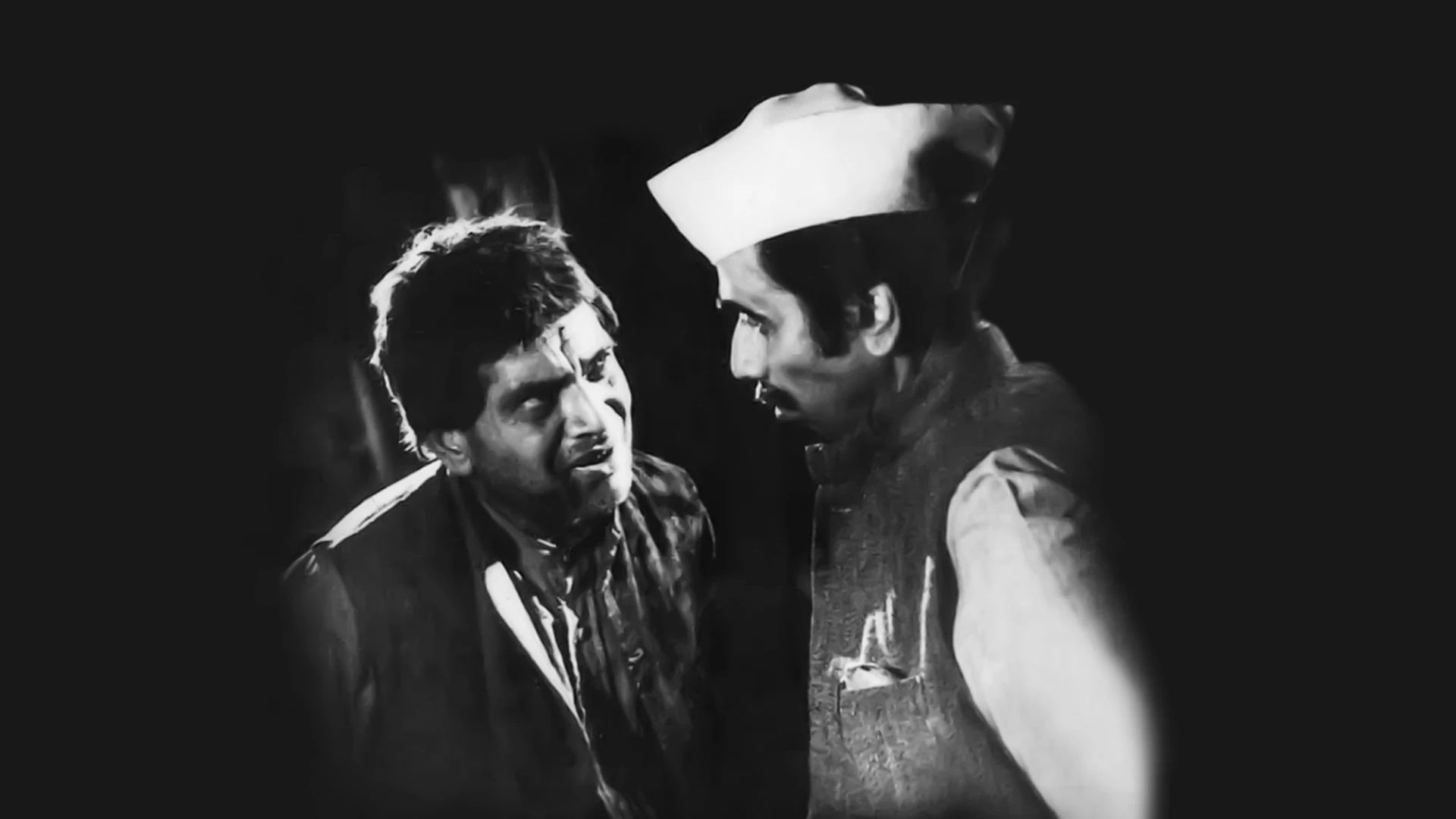This appears to be a black-and-white still from a film, showing two men in an intense conversation. The man on the left has an unkempt appearance, while the man on the right wears a Gandhi cap, possibly indicating a political or authoritative role. The dramatic lighting and expressions suggest a serious or emotional moment in the scene.