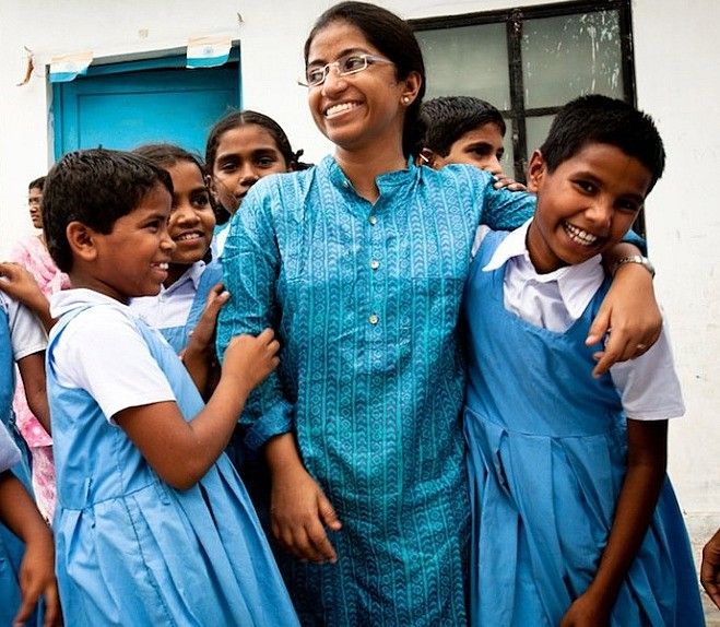 A smiling woman wearing glasses and a blue traditional outfit is surrounded by a group of young schoolgirls dressed in blue uniforms. The girls appear happy and are playfully interacting with the woman, who seems to be a teacher or mentor. The background features a school building with a blue door and window. The image captures a moment of joy and warmth in an educational setting.