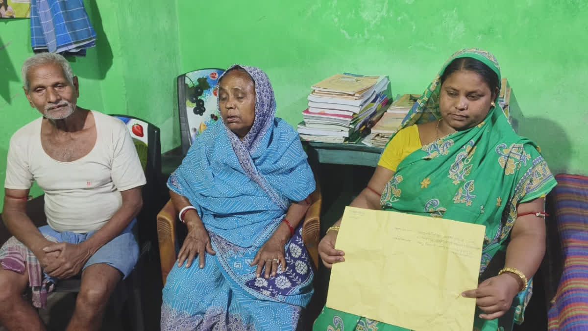 "A family sitting in a room with green walls, looking distressed. An elderly man and woman sit on chairs, while a younger woman in a green saree holds a yellow document. A table stacked with books and papers is visible in the background."