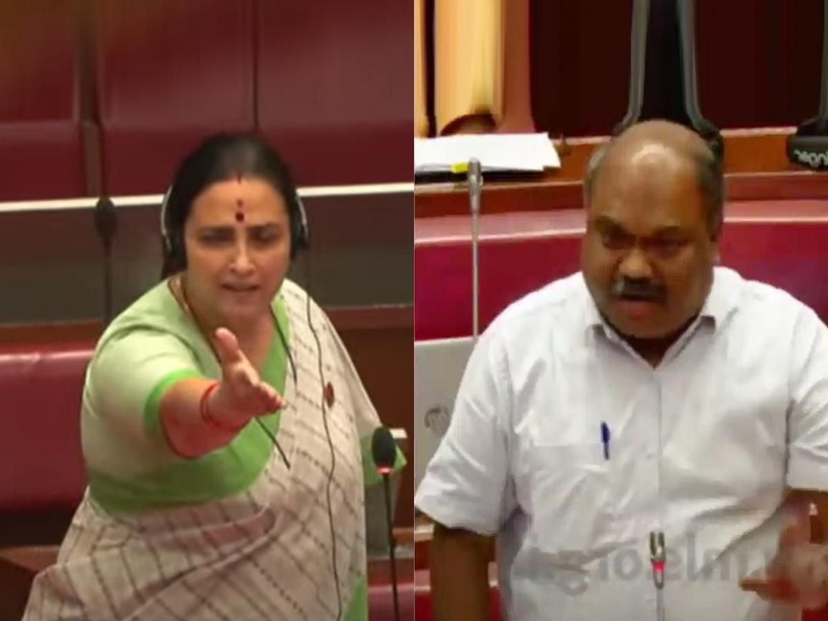This image appears to show two individuals engaged in a heated discussion or argument inside a legislative assembly or parliamentary setting. The woman on the left, dressed in a green saree, is extending her hand while speaking, and the man on the right, dressed in a white shirt, appears to be responding assertively. The background features red seats and microphones, suggesting a formal debate or political discussion.