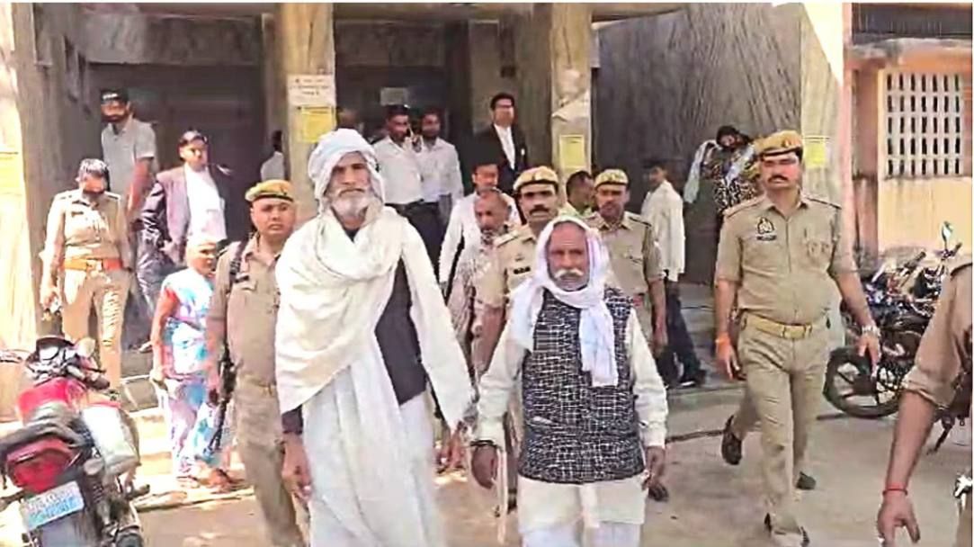 Two elderly men in traditional attire are escorted by police officers outside a courthouse. Several uniformed officers, lawyers, and bystanders can be seen in the background, indicating a legal proceeding or judicial action.