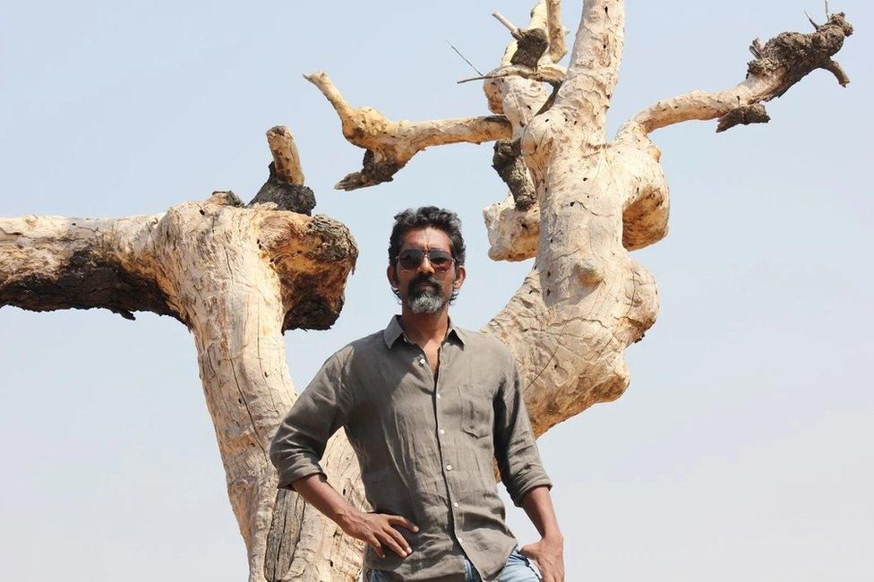 A man with a beard and sunglasses, wearing a casual gray shirt, stands confidently in front of a large, dried, and twisted tree under a clear blue sky. The background features a stark, barren landscape.