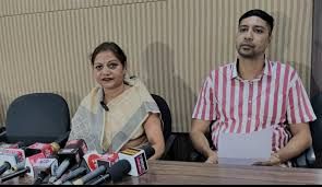 "A woman in a saree and a man in a striped shirt sitting at a press conference table with multiple microphones in front of them. The background features a beige panel wall, and they appear to be addressing the media."