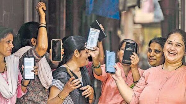 The image shows a group of women holding up their smartphones and smiling. They appear to be in a community or market area, and their expressions suggest excitement or happiness, possibly due to receiving some important news or benefits on their phones. The image conveys a sense of empowerment, digital access, or financial inclusion. Let me know if you need a detailed description or analysis!
