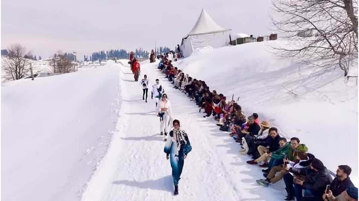 A unique fashion show is taking place on a snow-covered path in a scenic winter landscape. Models are confidently walking down the snowy runway, showcasing stylish outfits, while a large audience sits on both sides of the path, watching and capturing the moment on their phones. A white tent is visible in the background, along with snow-covered hills and a line of trees. The setting suggests a high-altitude location, possibly a famous winter tourist destination.