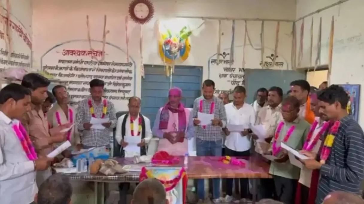 This image depicts a group of Indian men gathered in a room, seemingly taking an oath or participating in a formal ceremony. Many of them are wearing garlands, and they are holding papers while reading aloud. The room is decorated with banners and wall writings in Hindi or Marathi, suggesting a community or organizational event. Let me know if you need a more detailed description or specific context!
