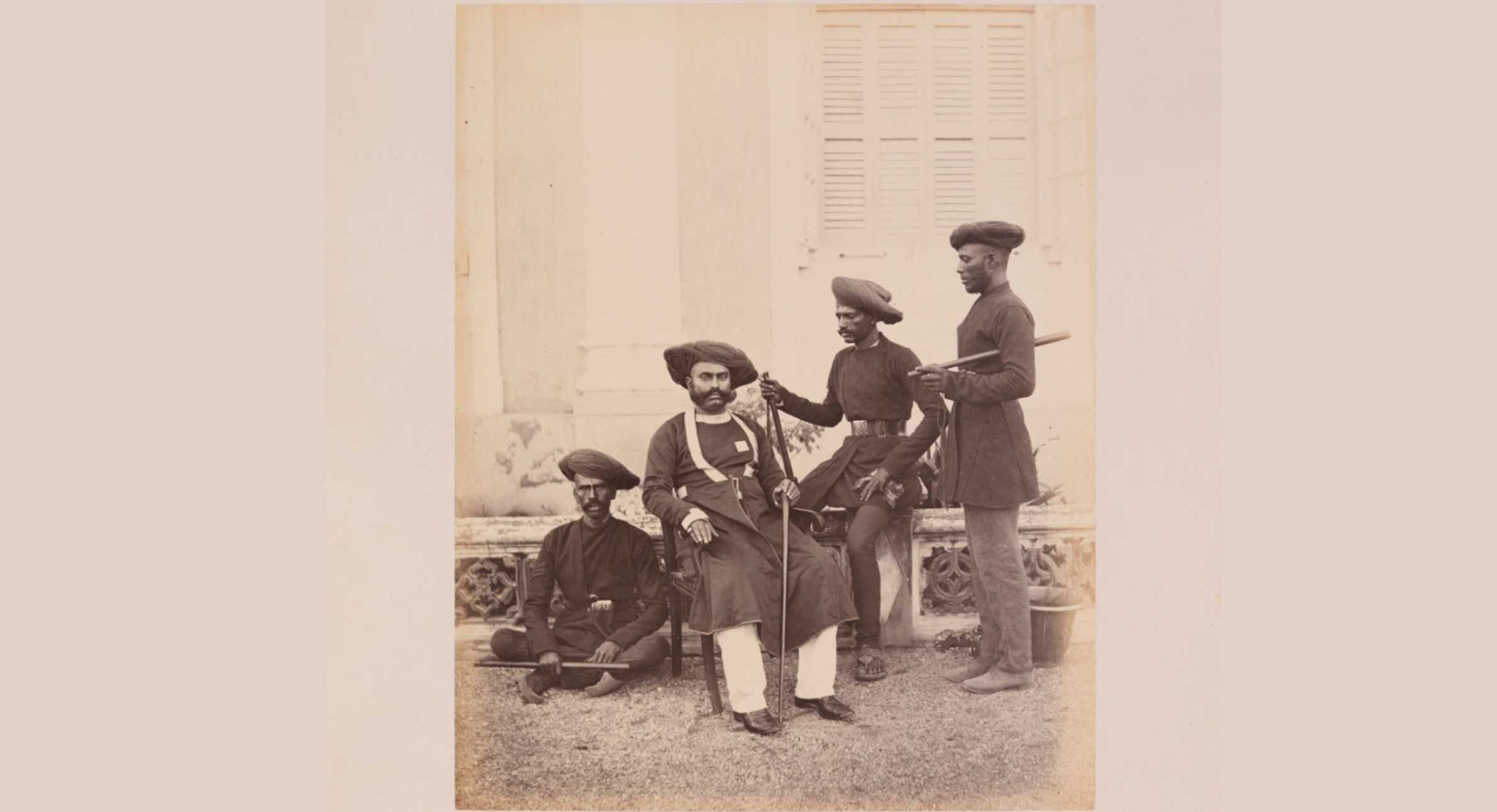 A historical sepia-toned photograph depicting a seated man in traditional Indian attire, surrounded by three standing men and one sitting on the ground. The men are dressed in dark tunics, turbans, and belts, with some holding weapons or batons. The backdrop features a colonial-era building with shuttered windows. The image likely represents a group of Indian guards or officials from the British colonial period in India.