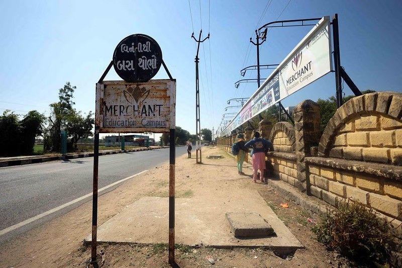 A rusted metal signboard on the side of a road reads "MERCHANT Education Campus" in English, with Gujarati text above it. The sign appears worn out, with peeling paint and rust. Behind the sign, a long yellow brick wall runs parallel to the road, adorned with banners displaying the "MERCHANT Education Campus" name. A group of students, dressed in colorful traditional attire, walk along the unpaved sidewalk beside the wall. Utility poles and power lines stretch down the road under a bright blue sky.