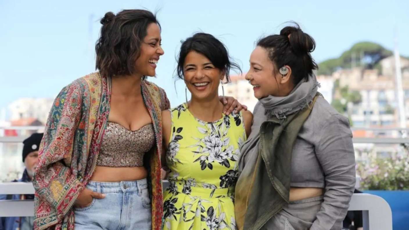 A photograph of three women laughing and embracing each other at an outdoor event, possibly a film festival. The woman on the left is wearing a glittery crop top, denim shorts, and a colorful bohemian-style jacket. The woman in the center is dressed in a bright yellow floral dress. The woman on the right is wearing a grey outfit with a scarf wrapped around her neck and has an earpiece in her ear. They appear joyful and are looking at each other, with a blurred background of a scenic outdoor setting, possibly near a waterfront or a cityscape.