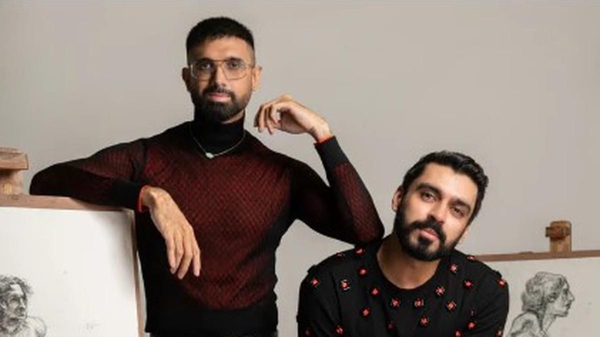 Two men pose stylishly in front of a neutral background, surrounded by artistic sketches on easels. The man on the left, wearing glasses and a black-red gradient sweater, leans on the easel with a confident expression. The man on the right, dressed in a black sweater with small red patterns, sits slightly tilted with a relaxed demeanor. Their contemporary fashion and artistic setting suggest a creative or designer-related theme.