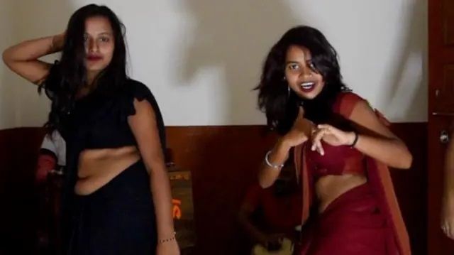 "Two young women in sarees dancing indoors with expressive poses. One is wearing a black saree, and the other is in a maroon saree. They appear to be enjoying the moment, with smiles and energetic movements. The background includes a wooden door and dim lighting."