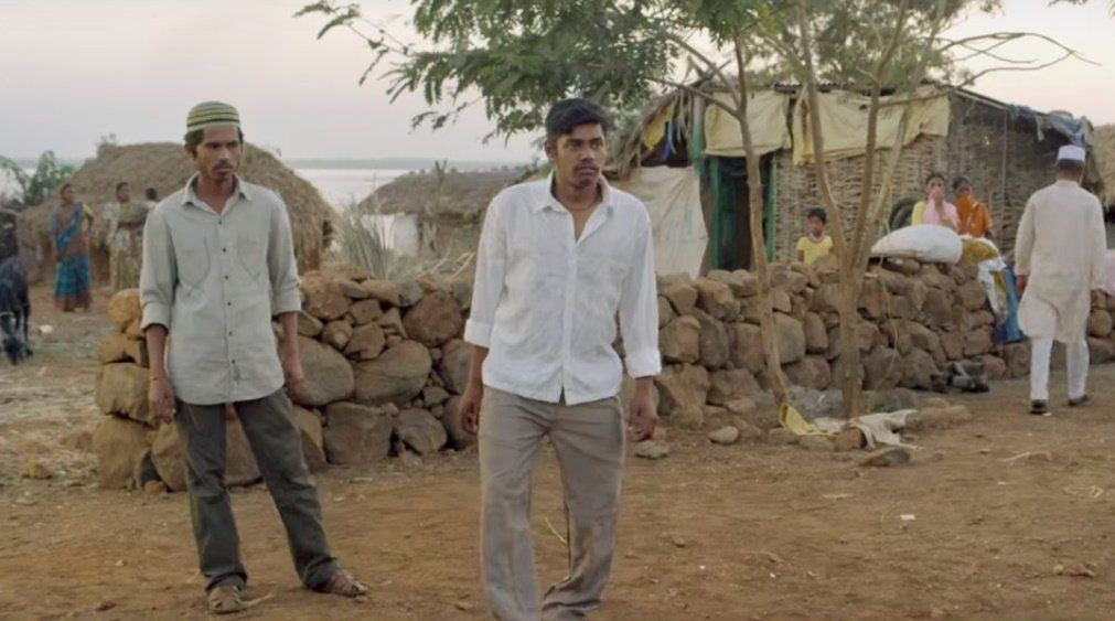 A rural setting with two men in the foreground, one wearing a white shirt and the other wearing a light green shirt with a cap, both appearing serious. In the background, there are stone walls, thatched huts, and makeshift houses made of bamboo and tarpaulin. Women and children are sitting near the houses, while another man in traditional white attire walks away. The environment has dry soil and scattered trees, suggesting a village near a water body visible in the distance.