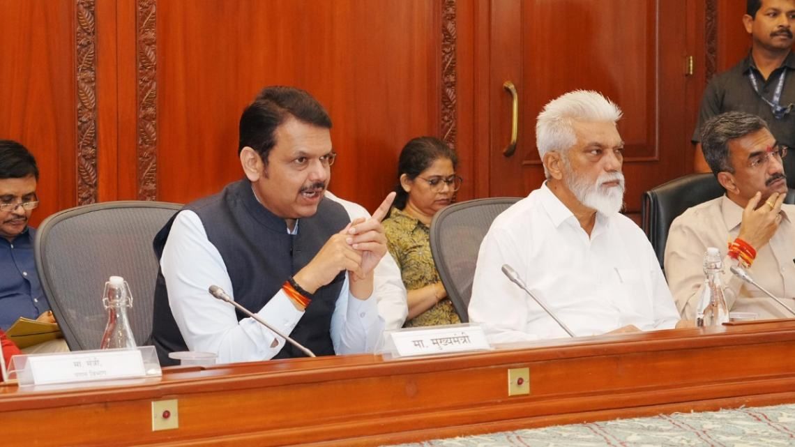 The image shows a formal meeting with officials seated at a wooden conference table. The central figure, wearing a dark vest over a white kurta, is speaking into a microphone with a serious expression and a hand gesture. To his right, an elderly man with a white beard in a white shirt listens attentively. Other officials, including a man in a beige uniform and a woman in glasses, are also present. The nameplate in front of the speaker reads "मा. मुख्यमंत्री" (Hon. Chief Minister), indicating a high-level government discussion. The setting features a polished wooden interior, microphones, and bottled water on the table.