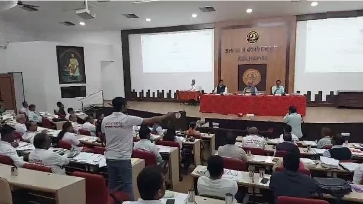 A conference hall at Shri J.J. University, Kolhapur, filled with attendees wearing white t-shirts with Marathi text "भ्रष्ट निवडणूक, निर्बंधित निवडणूक" (Corrupt Election, Restricted Election). The hall has a stage with three panelists seated behind a red table, addressing the audience. A participant in the crowd stands and gestures towards the stage, seemingly engaging in discussion. The room is well-lit, with a large screen displaying a presentation behind the panelists. The attendees appear to be actively participating in the session.