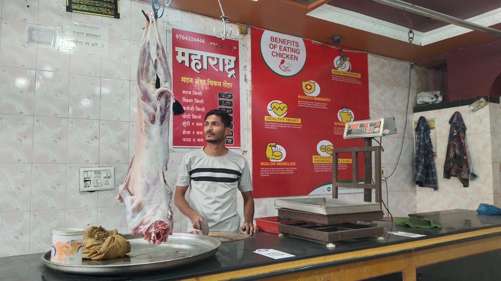 The image shows a butcher shop named "Maharashtra Matan & Chicken Center." A man wearing a light-colored T-shirt with black stripes is standing behind the counter, holding a knife near a chopping board. A large piece of raw meat is hanging from a hook beside him. The shop's interior has white tiled walls, and behind the man, there is a red sign displaying the "Benefits of Eating Chicken," including boosting immunity, improving heart health, and building muscles. An electronic weighing scale is placed on the counter, and some clothes are hanging on the wall in the background.