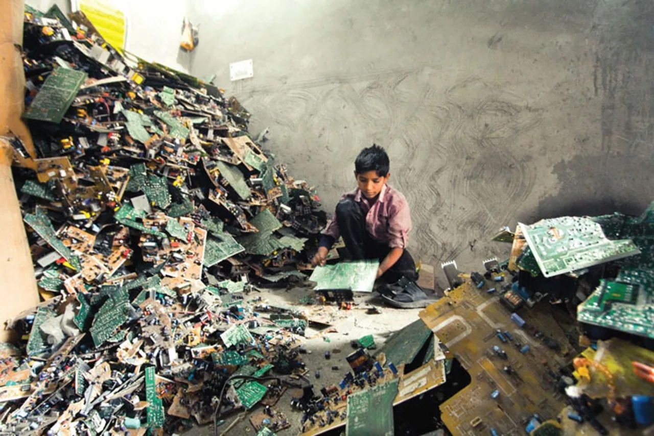"A young boy, wearing a worn-out shirt, sits on the floor of a dimly lit room filled with piles of discarded electronic circuit boards. He is handling a circuit board, surrounded by heaps of broken and dismantled electronic waste. The setting suggests hazardous child labor in the informal e-waste recycling sector, highlighting issues of environmental pollution and exploitation."