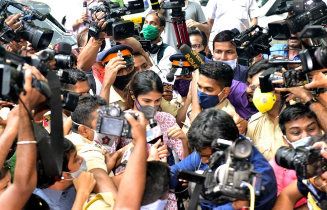 "A woman wearing a mask is surrounded by a dense crowd of media personnel and police officers. Multiple cameras, microphones, and reporters are seen pressing in while police officers attempt to escort her through the crowd."