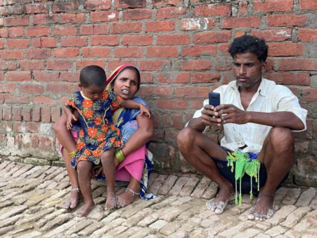 A rural family sits against a brick wall. A barefoot woman in traditional Indian attire holds a small child dressed in a colorful outfit, looking distressed. Beside her, a barefoot man in a worn-out shirt and shorts sits focused on a basic mobile phone, seemingly detached from the moment. The setting reflects rural life, digital divide, and possible struggles of daily survival.