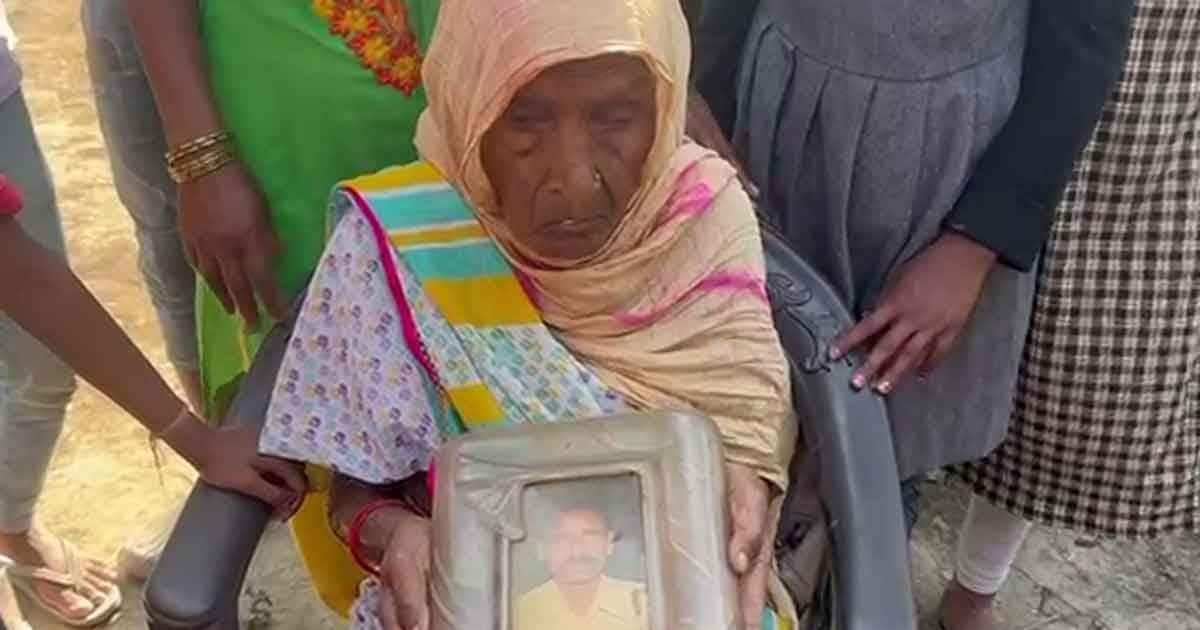 An elderly woman in traditional attire sits on a chair, holding a framed photograph of a man. She appears grief-stricken, surrounded by women and children, suggesting a moment of mourning or remembrance.