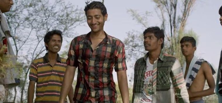 A young man in a checkered shirt smiles while walking, surrounded by a group of casually dressed men in an outdoor rural setting with trees in the background. The group appears engaged in conversation.