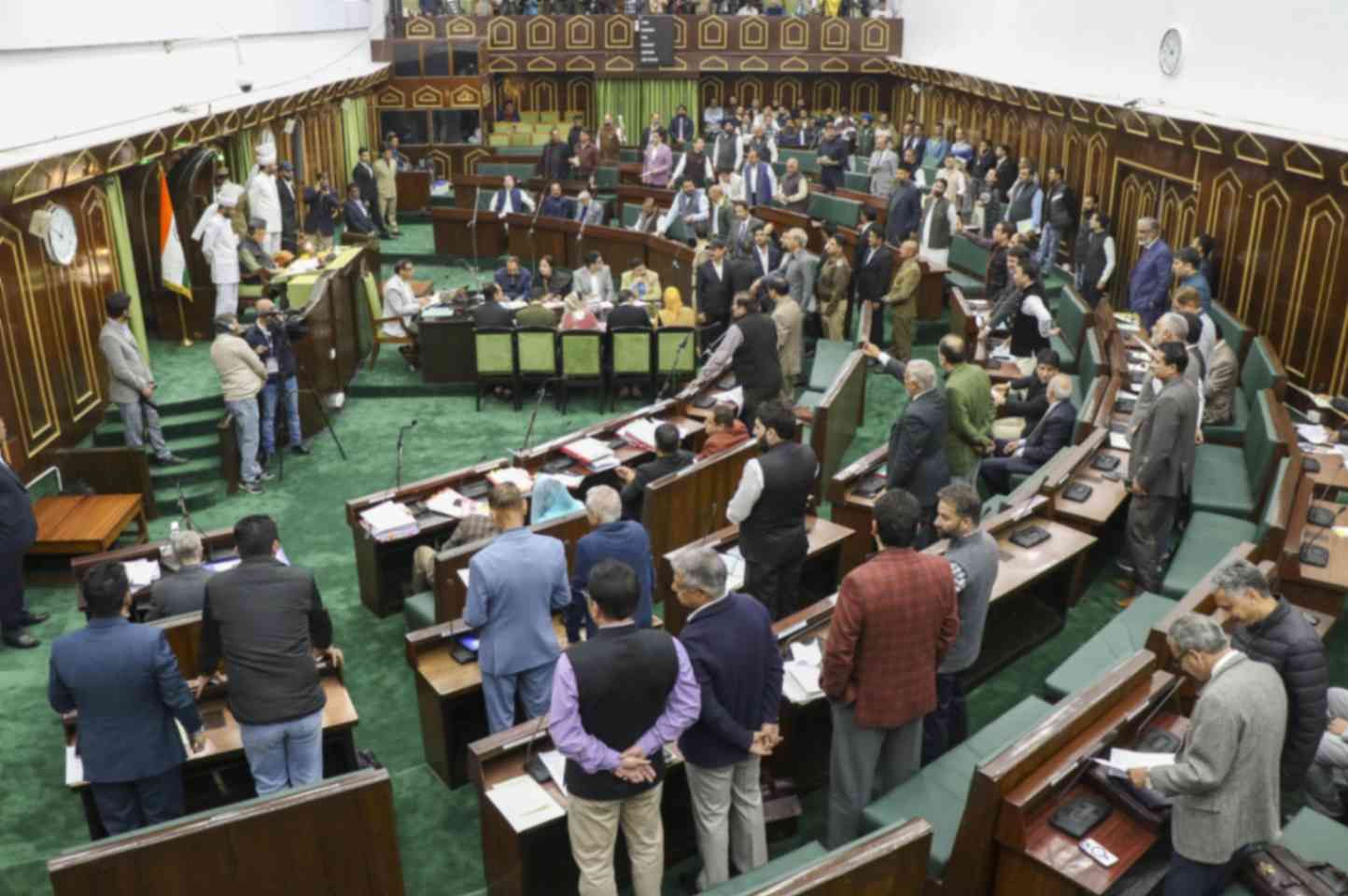 A legislative assembly hall filled with politicians and officials engaged in discussions. The hall has green carpets, wooden desks, and microphones. Some members are standing and conversing, while others are seated and reviewing documents. An Indian flag is visible near the speaker's podium, where officials are present. Journalists and camera operators are also capturing the proceedings.