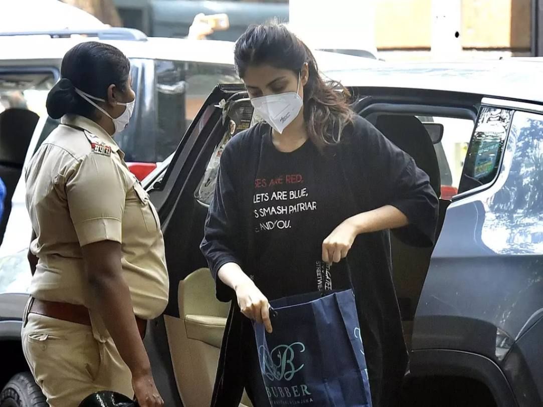 "A woman wearing a black outfit and a face mask steps out of a car holding a shopping bag, while a female police officer in uniform stands nearby. The background shows vehicles and an outdoor setting."