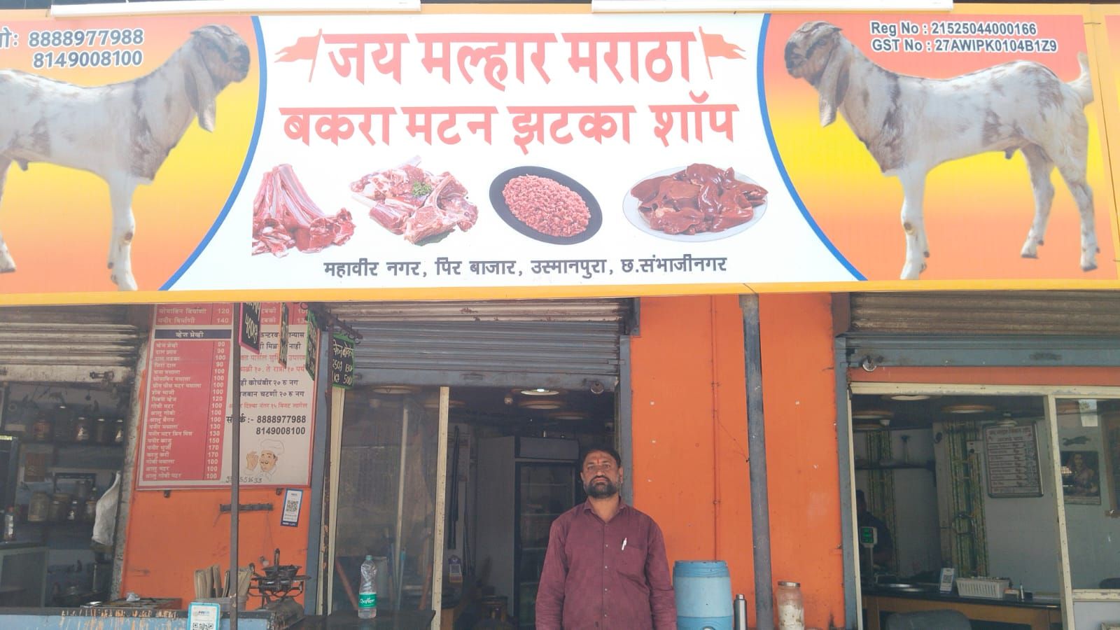 The image shows the exterior of a butcher shop named "Jai Malhar Maratha Bakra Mutton Jhatka Shop." The shop has a bright orange signboard featuring images of goats and different cuts of mutton, including ribs, minced meat, and liver. The location details on the sign indicate that the shop is in Mahavir Nagar, Pir Bazaar, Usmanpura, Chhatrapati Sambhajinagar. Two contact numbers and a GST registration number are also displayed. A man wearing a maroon shirt stands in front of the shop, looking at the camera. The shop's interior is partially visible, showing weighing scales, utensils, and a price board.