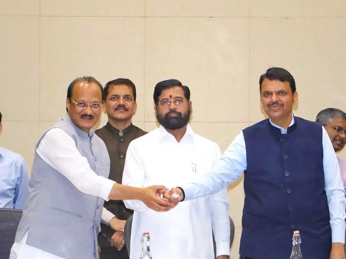 The image shows three prominent politicians shaking hands in a formal setting. The central figure, wearing a white kurta, stands between two other leaders who are shaking hands, symbolizing an agreement or alliance. The background consists of officials and a conference room setting, suggesting a political meeting or announcement. The expressions appear formal and strategic, indicating a significant political development. Let me know if you need a detailed description!
