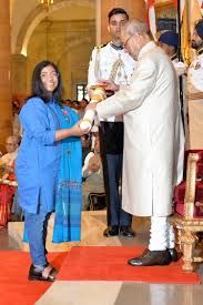 Sunitha Krishnan, an Indian social activist, is seen receiving a prestigious award from the President of India at an official ceremony. She is wearing a blue outfit and smiling as she accepts the honor. The President, dressed in traditional Indian attire, hands over the award while uniformed officers stand in the background. The event takes place in a grand hall with red carpeting and golden decor, symbolizing the significance of the occasion.