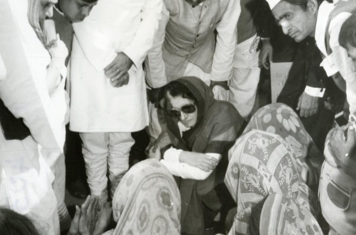 A black-and-white photograph captures a woman wearing sunglasses and a headscarf, sitting on the ground with folded arms, engaging with a group of women dressed in traditional attire. She is surrounded by men in formal and traditional clothing, indicating a moment of serious discussion or condolence. The image conveys a sense of empathy and social engagement.