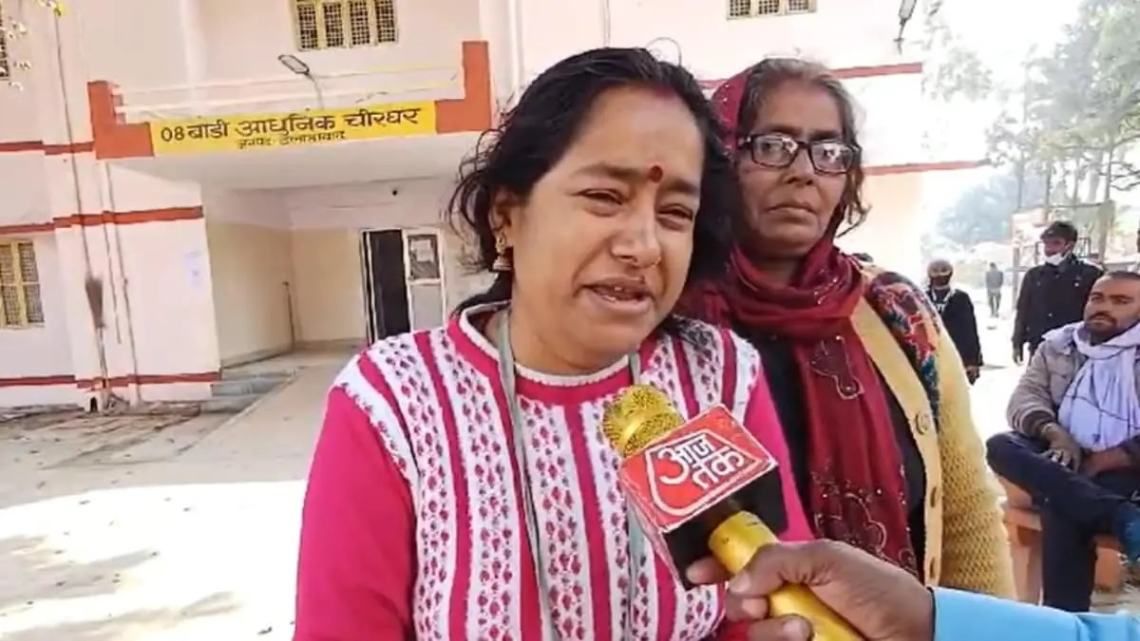 "A distressed woman in a pink and white sweater is being interviewed by a journalist holding a microphone with the 'Aaj Tak' logo. Another woman wearing glasses and a red scarf stands behind her. The background shows a building with a Hindi sign reading '08 बाड़ी आधुनिक चौखर,' along with several people sitting and standing."