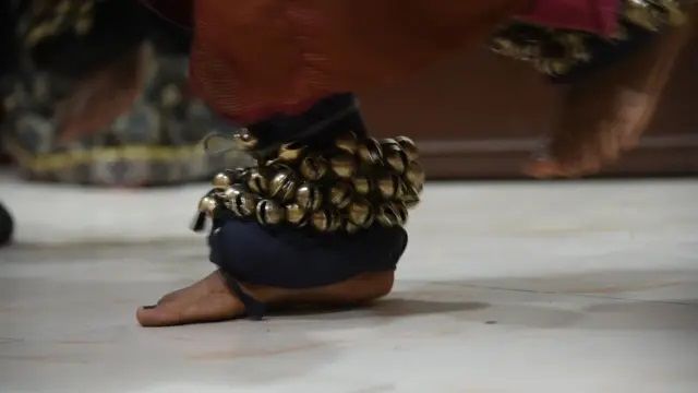 "A close-up of a dancer’s feet adorned with traditional ghungroos (ankle bells) in motion. The dancer is barefoot, and the bells create a rhythmic visual as they move on a polished floor. The background is slightly blurred, highlighting the intricate details of the ghungroos and the dancer's footwork."