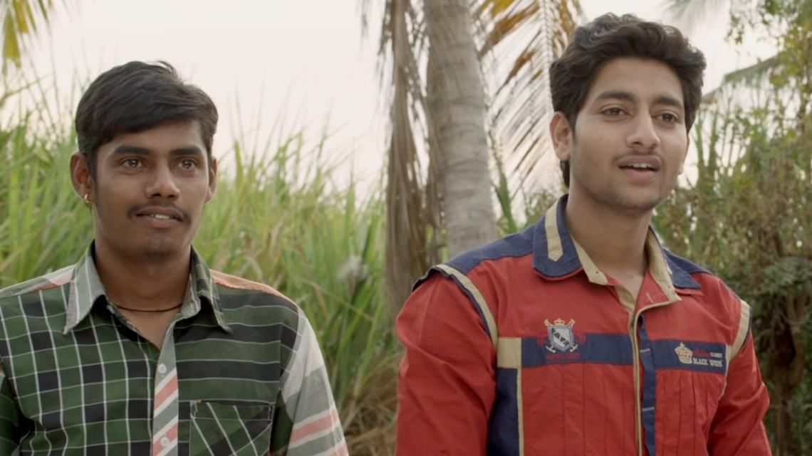 Two young men are sitting outdoors in a rural setting, surrounded by lush green sugarcane fields and palm trees. The man on the left is wearing a dark green checkered shirt and has a slight smile, while the man on the right is dressed in a red and blue jacket with a logo on the chest, speaking with an enthusiastic expression. The natural background suggests a village or agricultural area.