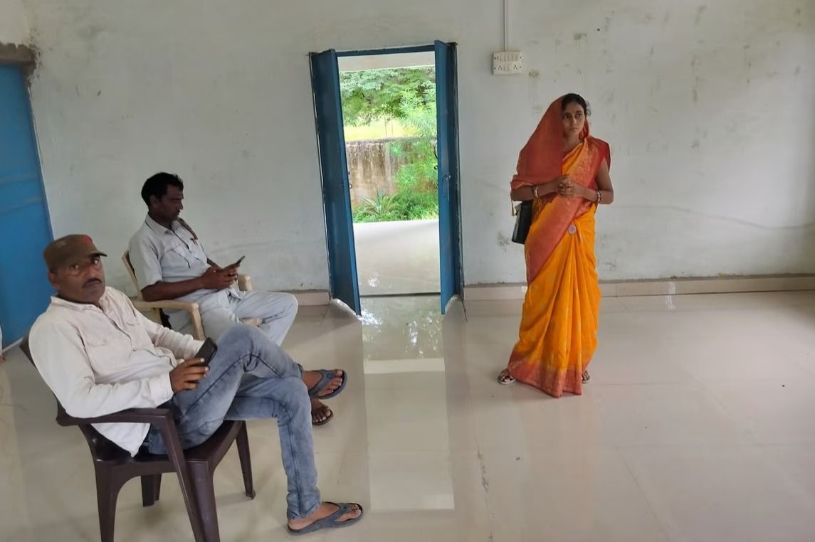 The image shows a woman in a yellow saree standing in a simple room with a tiled floor, while two men are seated on plastic chairs. One of the men is looking at the camera, while the other is using his phone. The background features an open door leading to a green outdoor area. The setting appears to be a rural or semi-urban community space. Let me know if you need any specific details!
