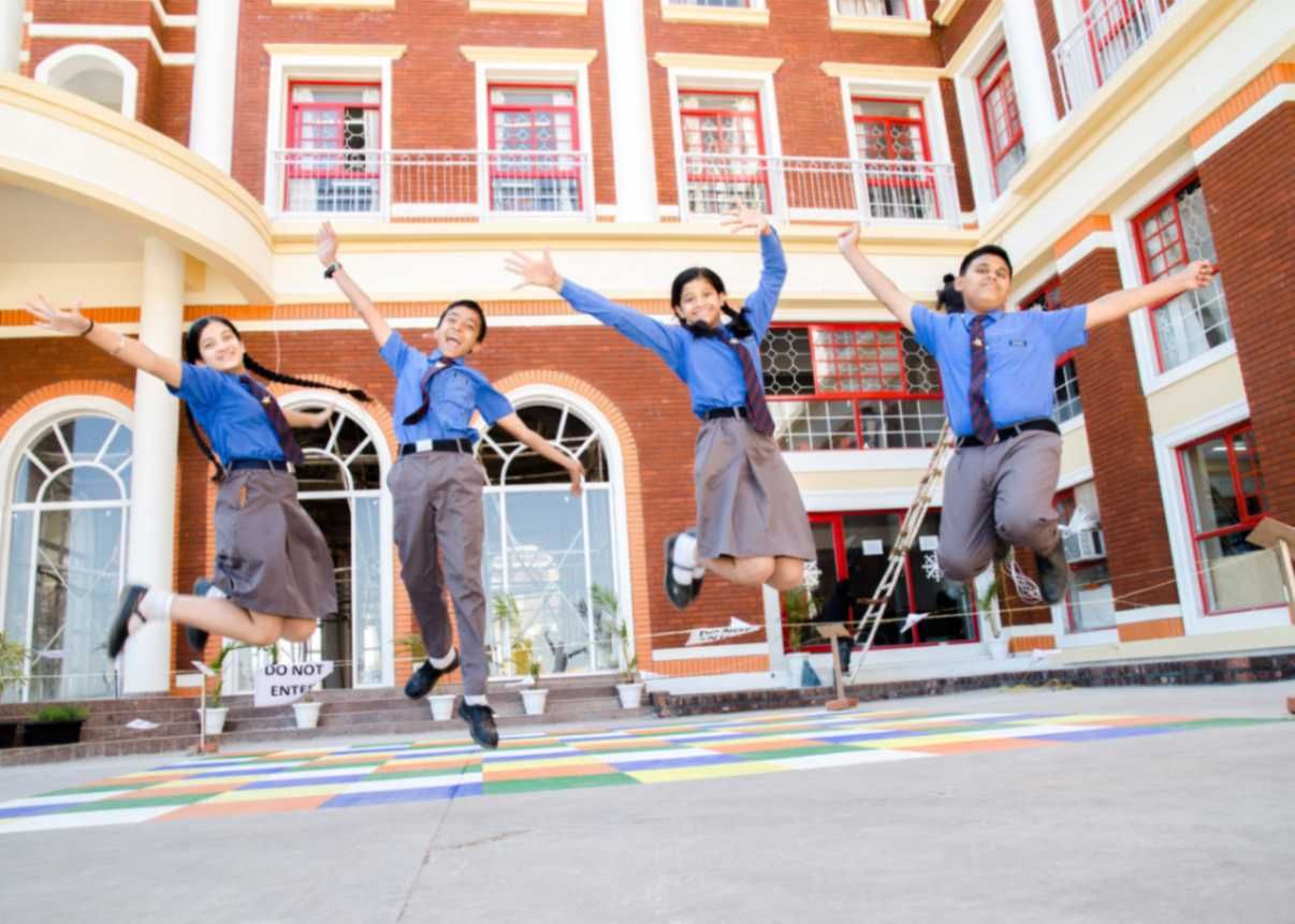 The image shows four school children in blue uniforms joyfully jumping in the air in front of a grand school building with a red and cream-colored facade. The students, two boys and two girls, appear excited and happy, possibly celebrating an achievement or enjoying a break. The building features large arched windows, white pillars, and a decorative entrance, giving it a prestigious appearance. The vibrant school courtyard is decorated with colorful patterns on the ground. A sign reading "DO NOT ENTER" is visible in the background.