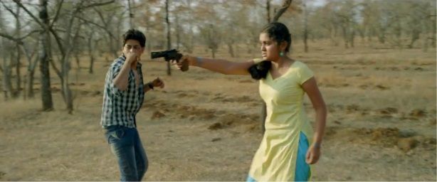 A young woman in a yellow and blue outfit points a handgun with determination in an open, dry landscape. Beside her, a young man in a checkered shirt looks startled, seemingly reacting to the tense situation. The background features sparse trees and dry terrain, adding to the dramatic intensity of the moment.