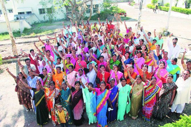 The image shows a large group of women, many wearing colorful sarees, gathered outdoors in a spirited pose with raised fists. Some men are also present in the background. The setting appears to be a community or rural area, possibly outside a building. The scene conveys a sense of unity, empowerment, and collective action. Let me know if you need any specific details!