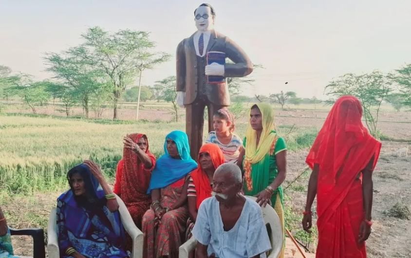 A group of rural men and women, dressed in traditional colorful attire, are seated and standing in an open field, engaged in conversation. Behind them stands a statue of Dr. B.R. Ambedkar, depicted in a suit holding the Indian