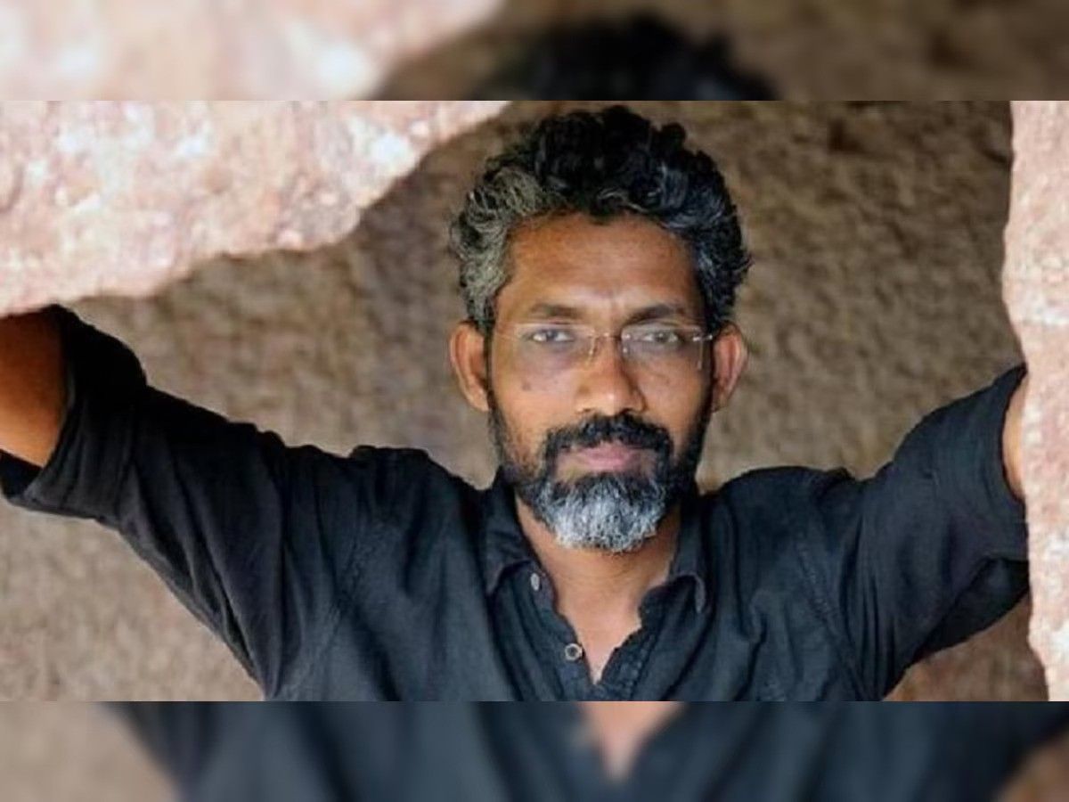 This is a photograph of a man with salt-and-pepper hair and a beard, wearing a black shirt and glasses. He is positioned between two rocky formations, with his arms resting on them. His expression is calm and serious, and the background consists of a rough, stone-like texture. The image has a blurred reflection effect at the top and bottom.