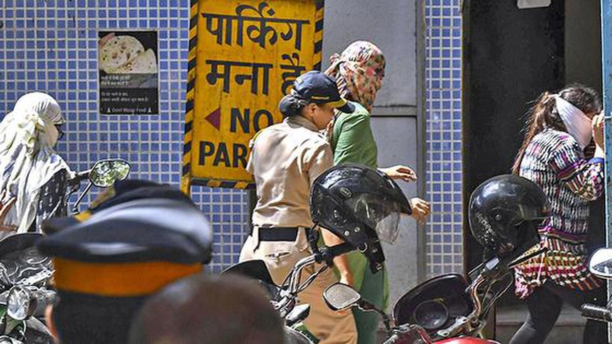 A police officer in a beige uniform is escorting a woman in a green outfit with her face covered by a patterned cloth into a building. Another woman, also covering her face, is seen entering the building. In the foreground, several police personnel and motorcycles with black helmets are visible. A yellow sign with Hindi and English text reads "पार्किंग मना है" (No Parking) on a tiled blue wall in the background, along with a food advertisement poster. The scene appears tense, suggesting a police-related action or investigation.