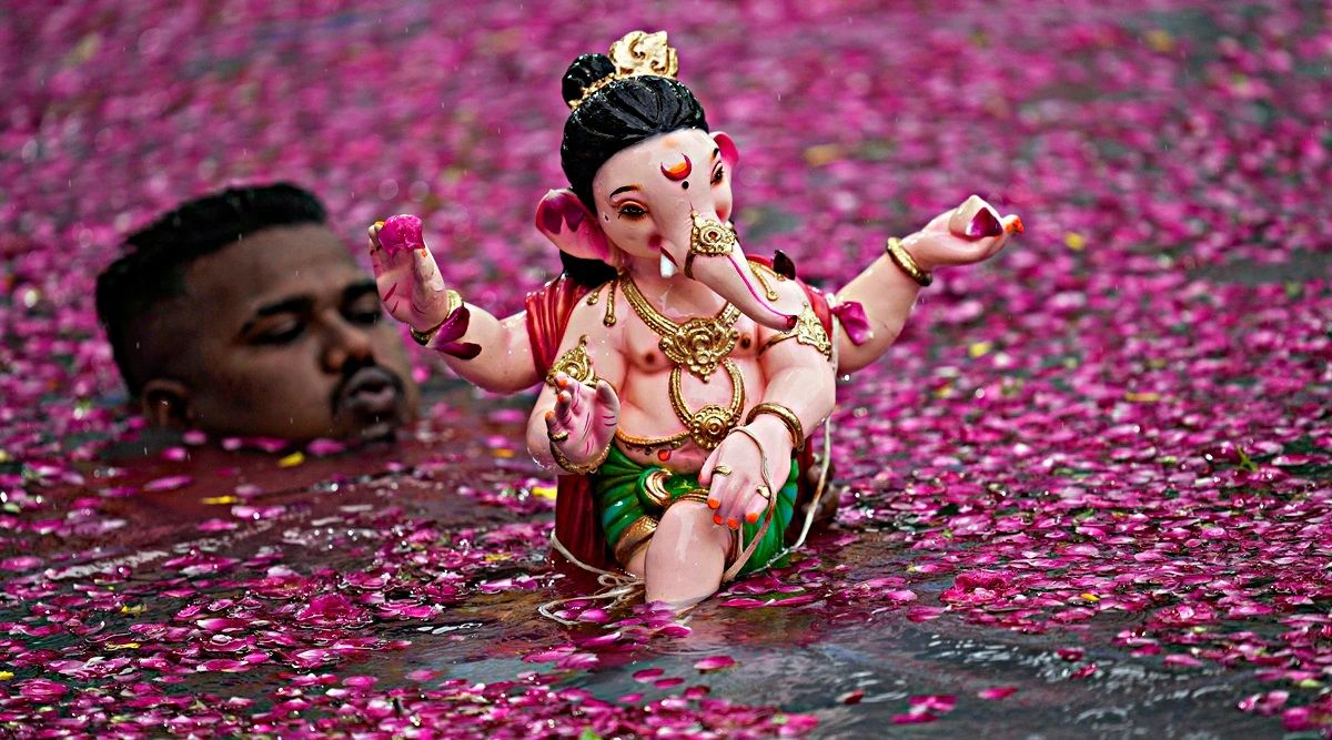 "A beautifully adorned idol of Lord Ganesha is seen floating on water covered with pink flower petals during an immersion ceremony. In the background, a man is partially submerged in the water, assisting with the visarjan (idol immersion) ritual. The image captures the vibrant and emotional essence of Ganesh Chaturthi celebrations."