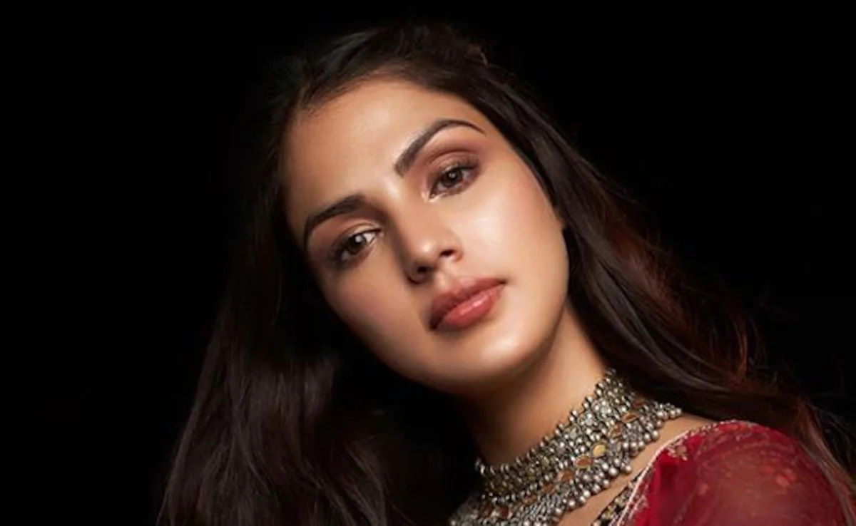 "A close-up portrait of a woman with long brown hair, wearing a red traditional outfit and an ornate silver necklace. She has a calm expression, with subtle makeup, against a dark background."