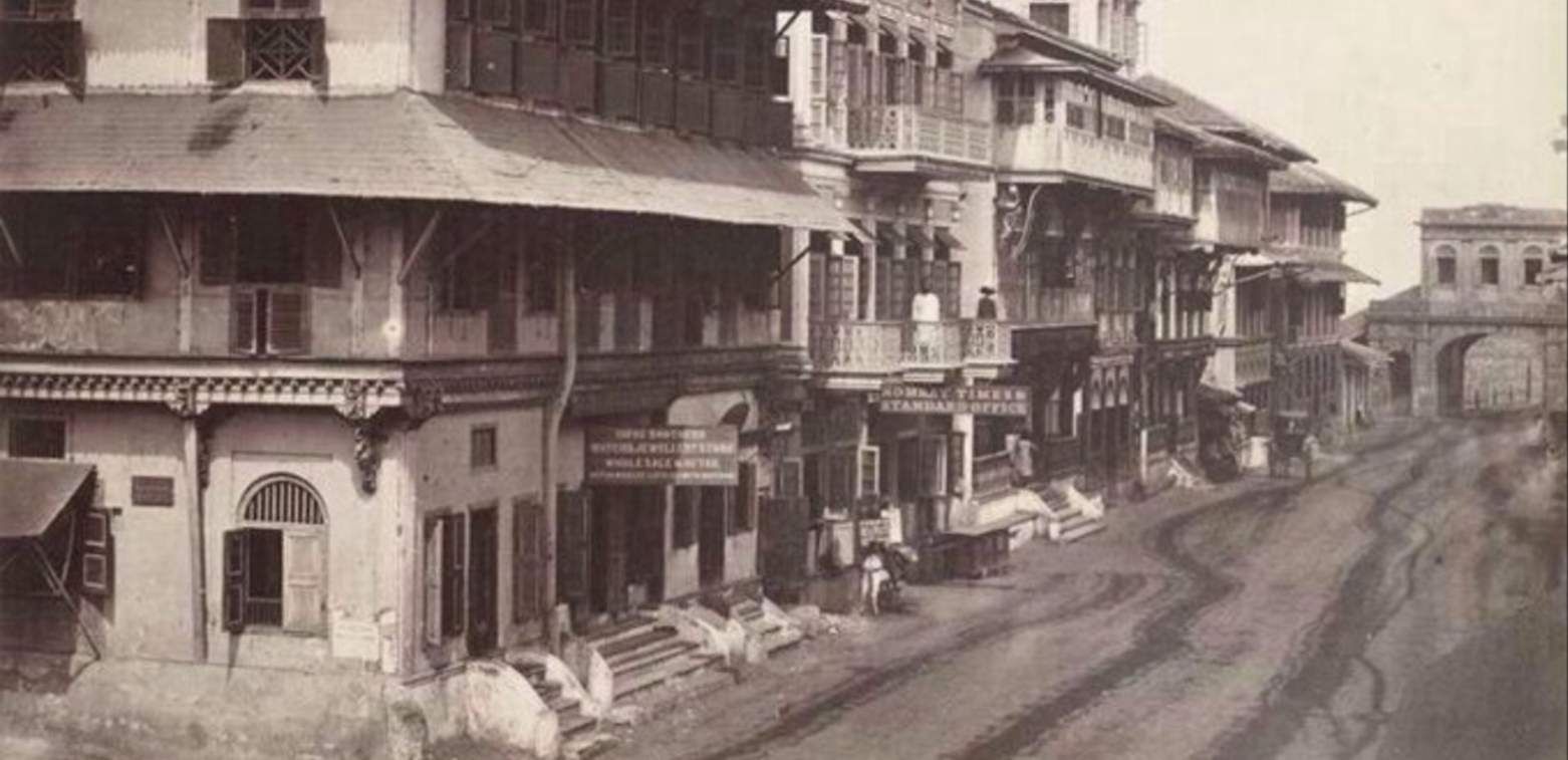 A historical black-and-white photograph of a street in colonial-era Bombay (now Mumbai), lined with multi-story wooden buildings featuring intricate balconies and signage. The street appears unpaved, with a few people visible on balconies and near shop entrances. A signboard for the "Bombay Times Standard Office" is prominently displayed, indicating the presence of a newspaper office. The background features an arched gateway, adding to the architectural charm of the period.
