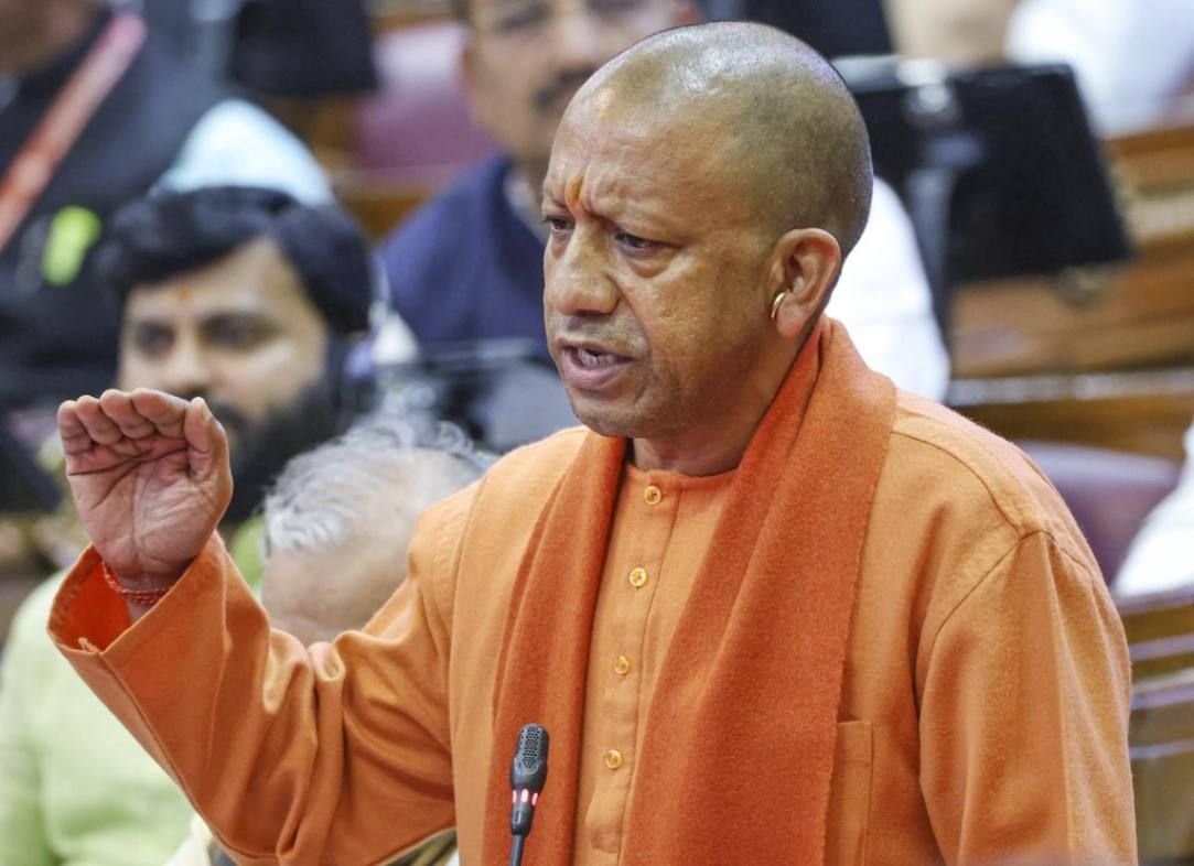 "A bald man wearing a saffron robe and scarf is speaking passionately in a legislative assembly. He is gesturing with his right hand while addressing the gathering. Several people are visible in the background, some wearing traditional Indian attire, while others are dressed in formal clothing."